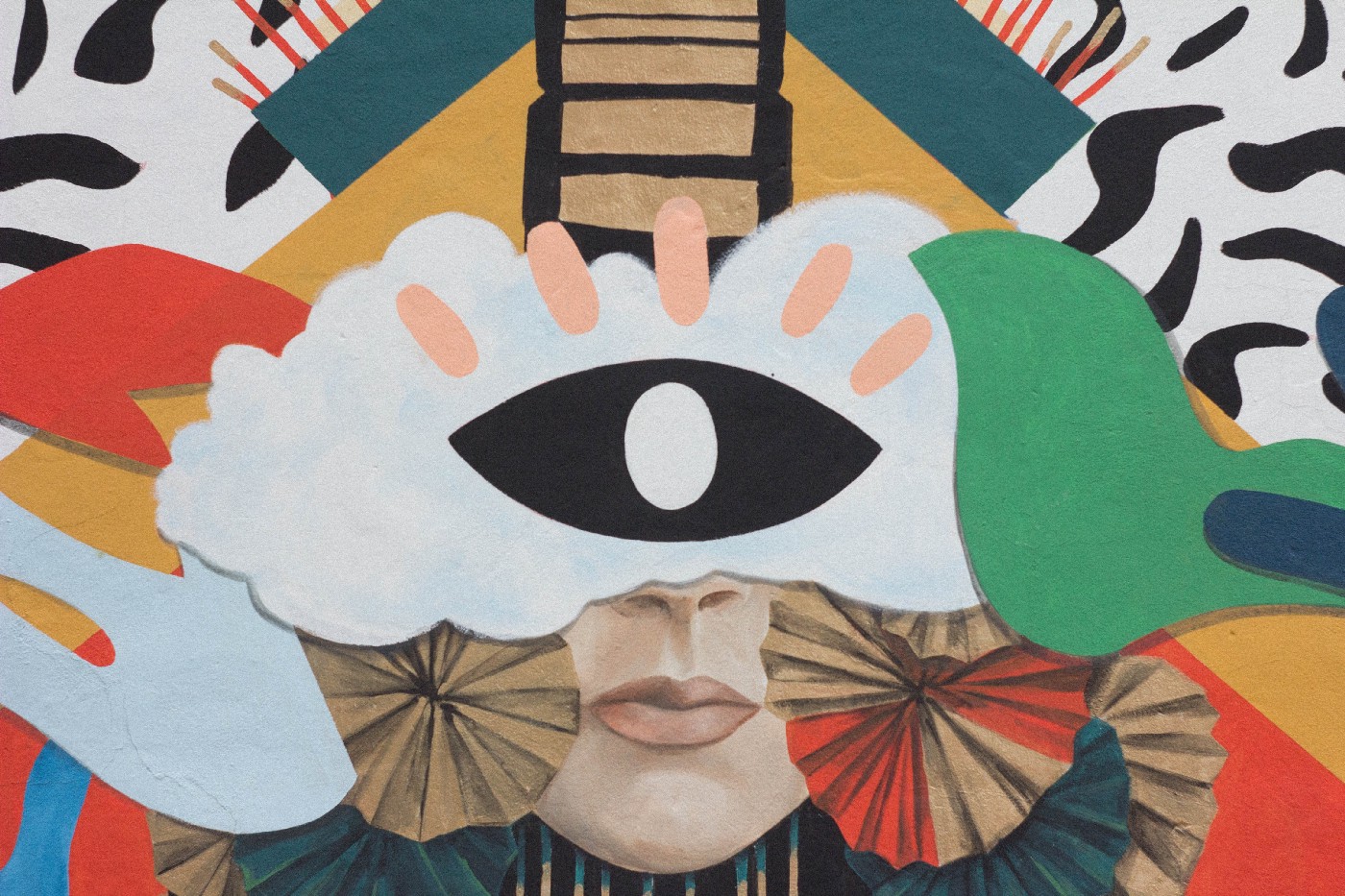 Part of a face surrounded by colourful images, looks like a wall-painting, with big black-and-white eye where the forehead would be if it weren’t behind what looks like a cloud