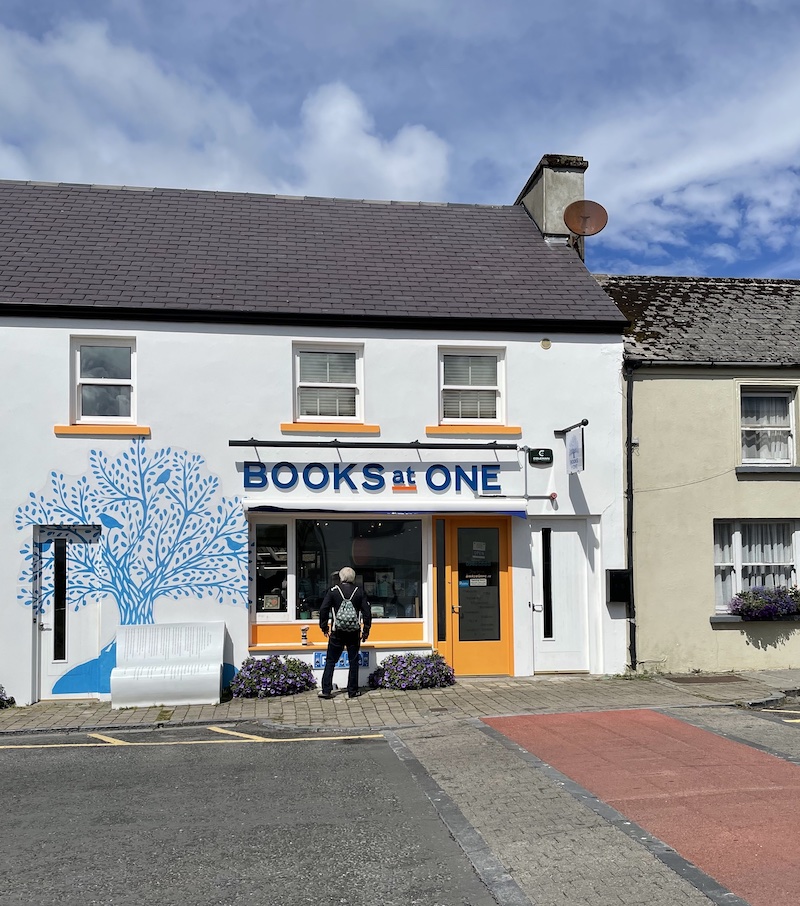 The front of Books at One bookshop in Louisburgh, County Mayo; the front wall is white with a stylized tree with birds painted on it in blue to the left of the shop display window, the shop door and window ledges are painted a mustardy yellow.